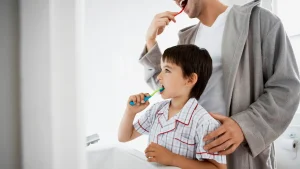 A father teaches his son good brushing habits, a key part of daily kids dental care to prevent cavities.